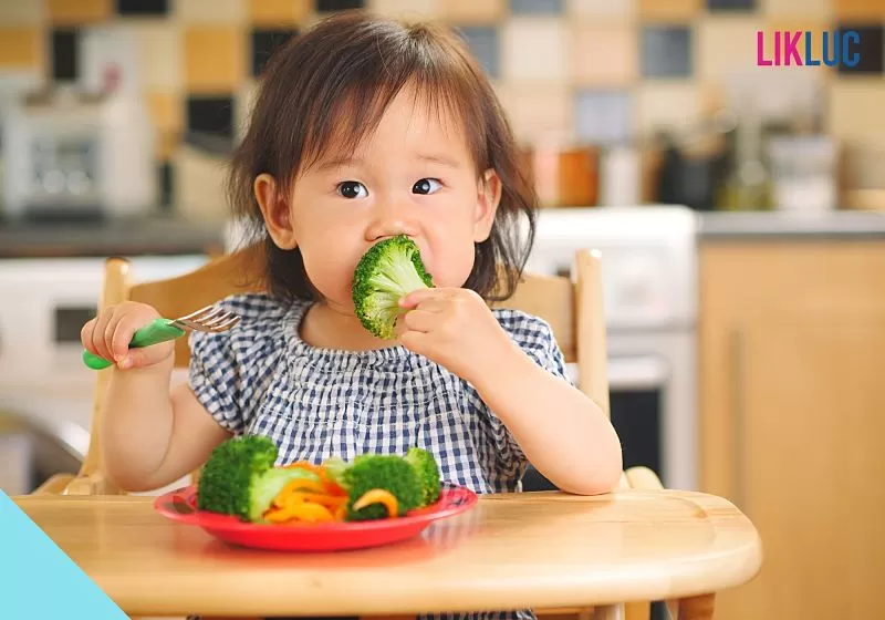 Bebê sentado em sua cadeira de comer com um prato com brócolis e cenoura