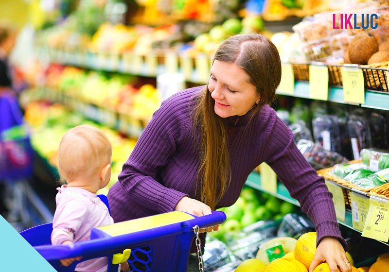 mulher em sessão de hortifruti de supermercado com carrinho de compras azul e bebê na cadeira do carrinho, olhando para a criança