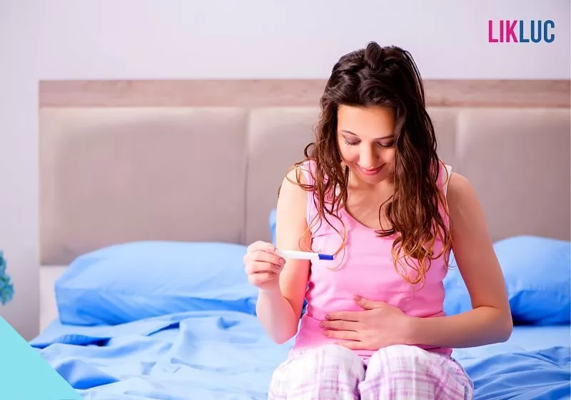 mulher sentada na cama segurando barriga com uma mao e teste de gravidez na outra
