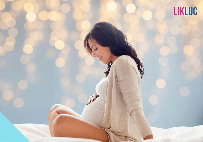 gestante sentada de lado com mao na barriga sorrindo e luzes brilhantes no fundo
