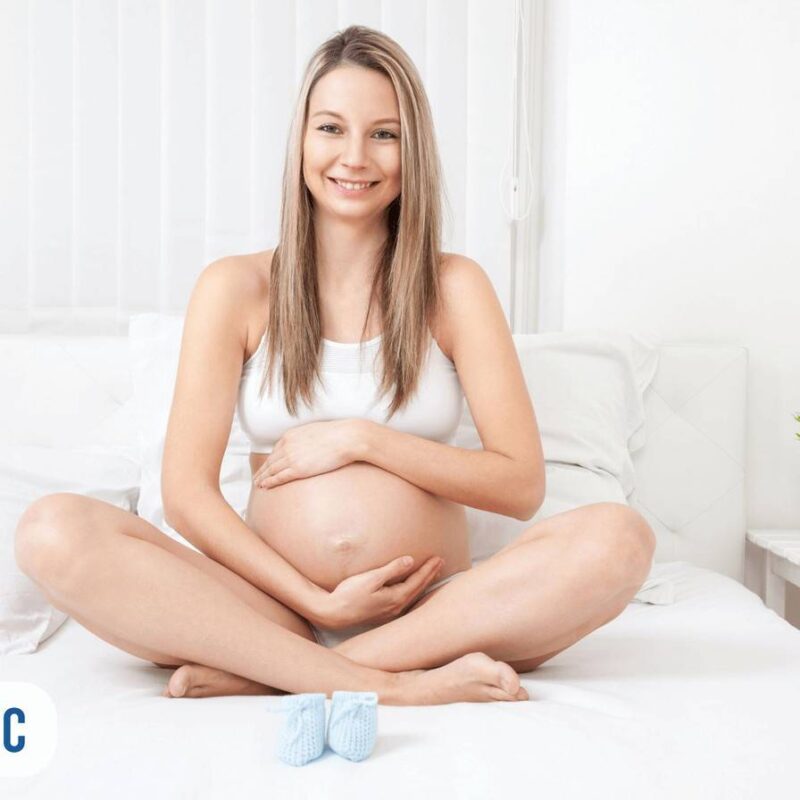 Gestante sentada com as pernas cruzadas segurando a barriga e na sua frente um par de sapatinhos de bebê