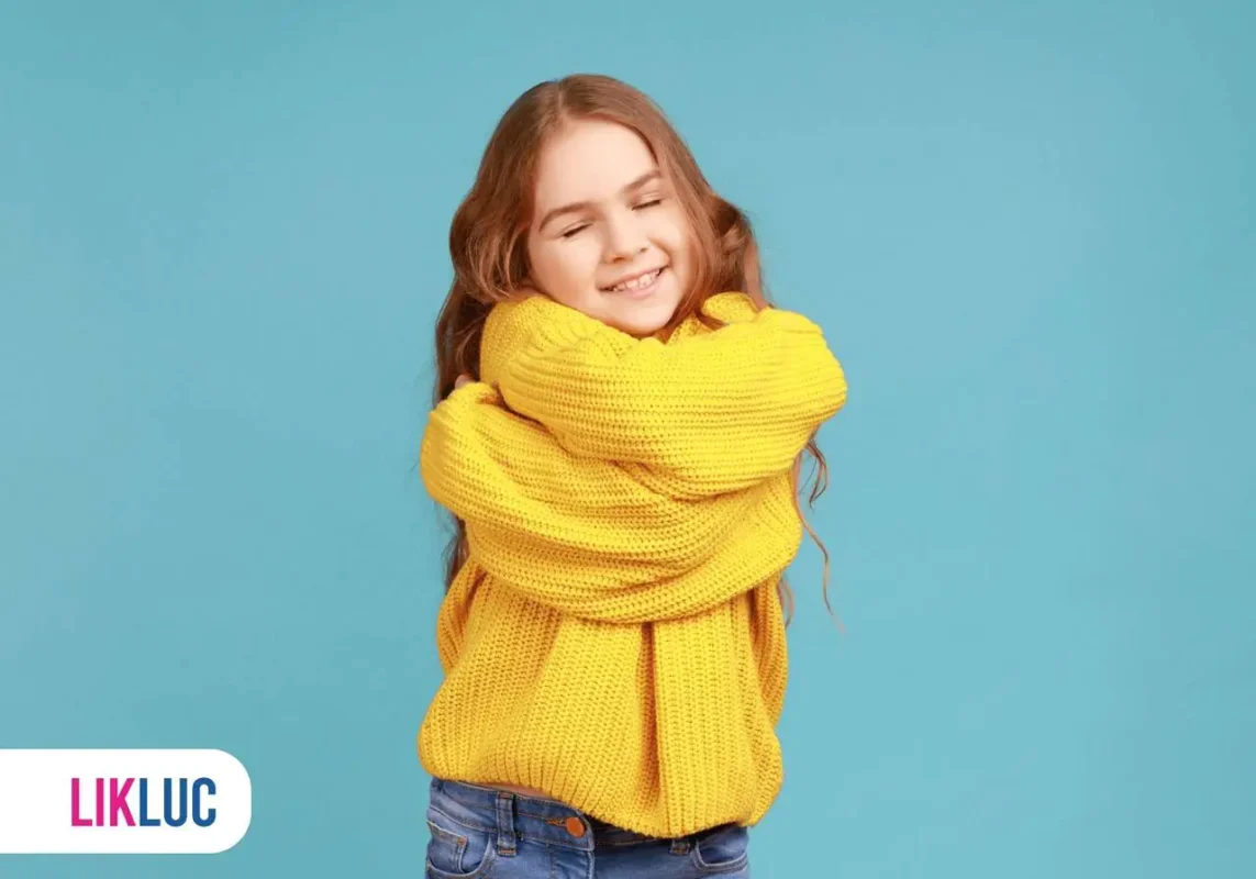 Criança se abraçando vestindo blusa amarela com calça jeans azul, ao fundo parede de cor azul claro