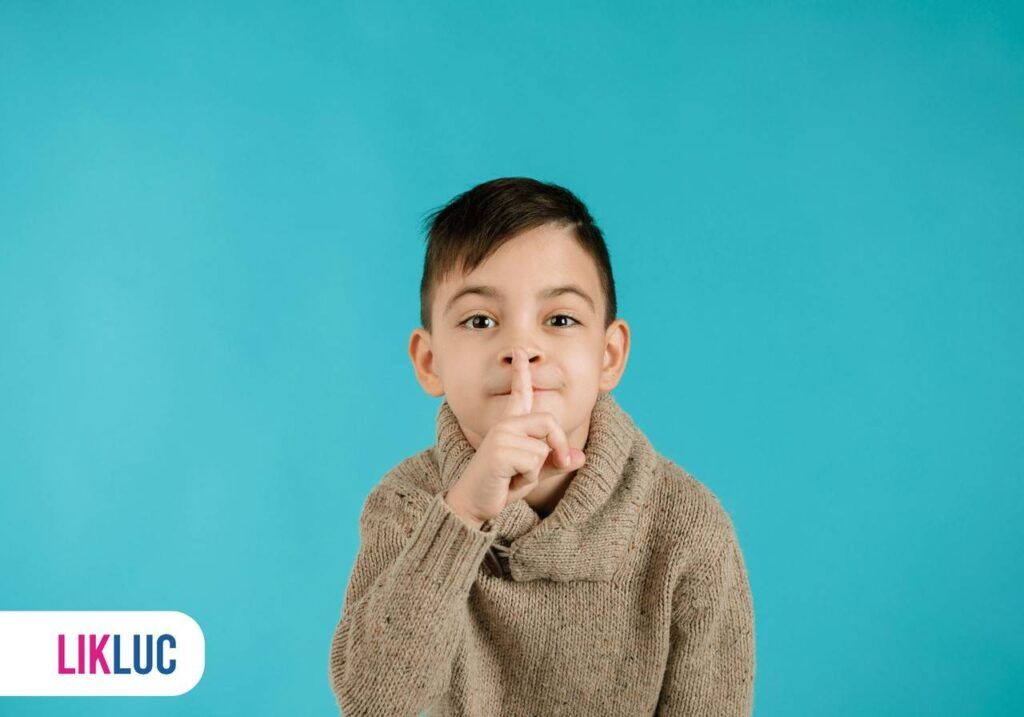 Criança usando blusa no tom marrom claro, fazendo gesto de silêncio e ao fundo azul claro