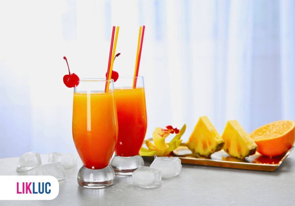 dois copos de vidro com sucos e canudo, decorados com fruta, ao lado, bandeja com frutas cortadas.