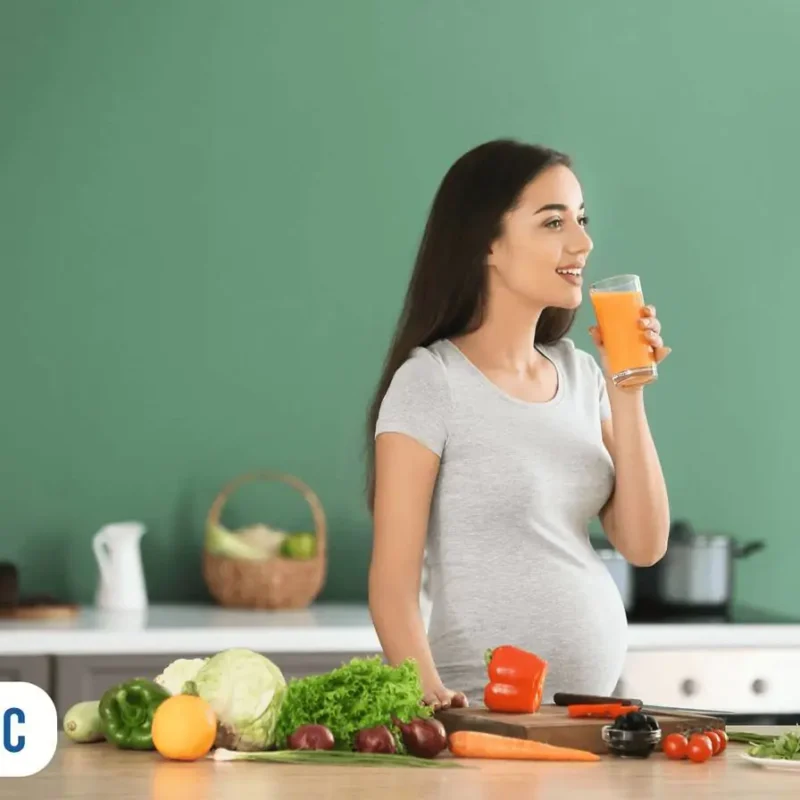 Mulher com copo de suco na mão, ao lado de mesa com tábua e alguns legumes cortados