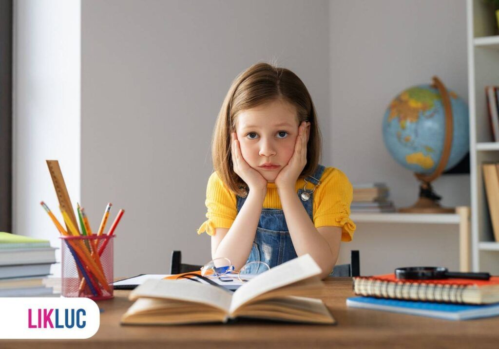 Criança em mesa com as duas mãos sobre o rosto, na mesa livros, lápis e caderno