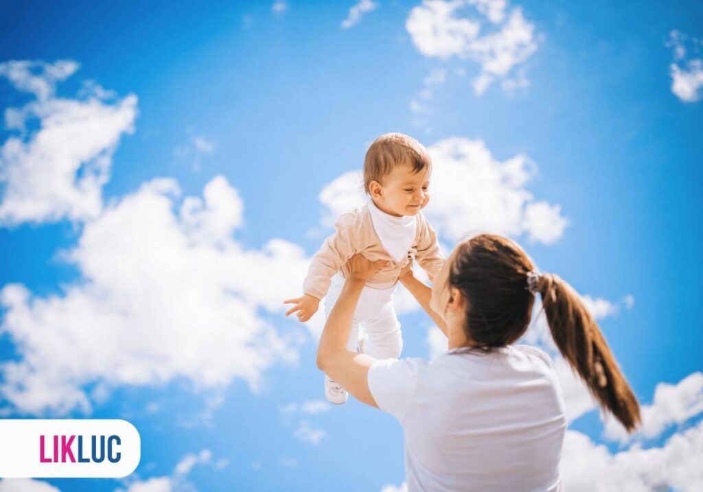 Mãe segurando bebê com os braços levantados, ambos utilizando roupas brancas, ao fundo, ceú azul com nuvens