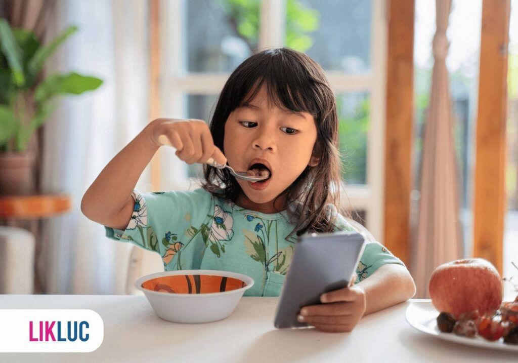 Criança em mesa, comendo com uma mão, segurando e observando celular