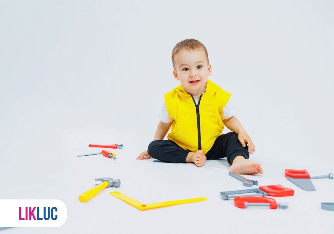 Criança sentada ao chão, usando colete amarelo e calça preta, com brinquedos espalhados que simulam ferramentas de construção