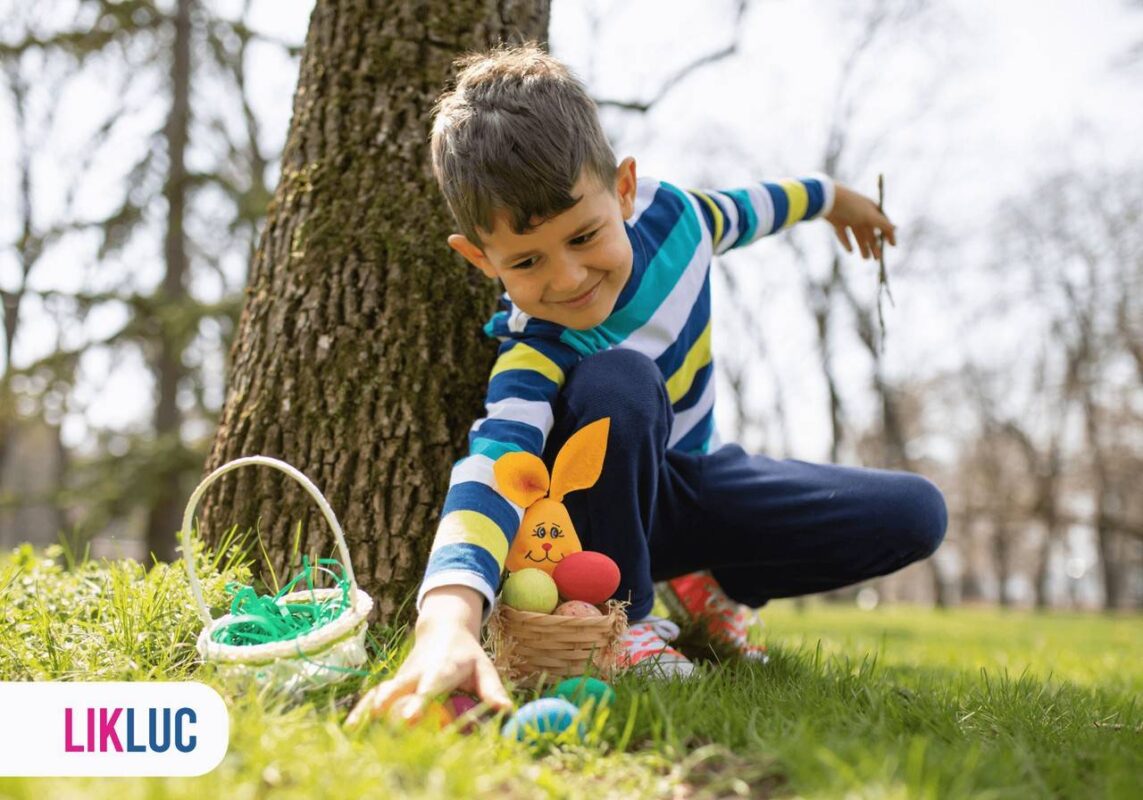 Criança em praça com árvores e grama, sentado e tentando alcançar cesta com óvos de páscoa