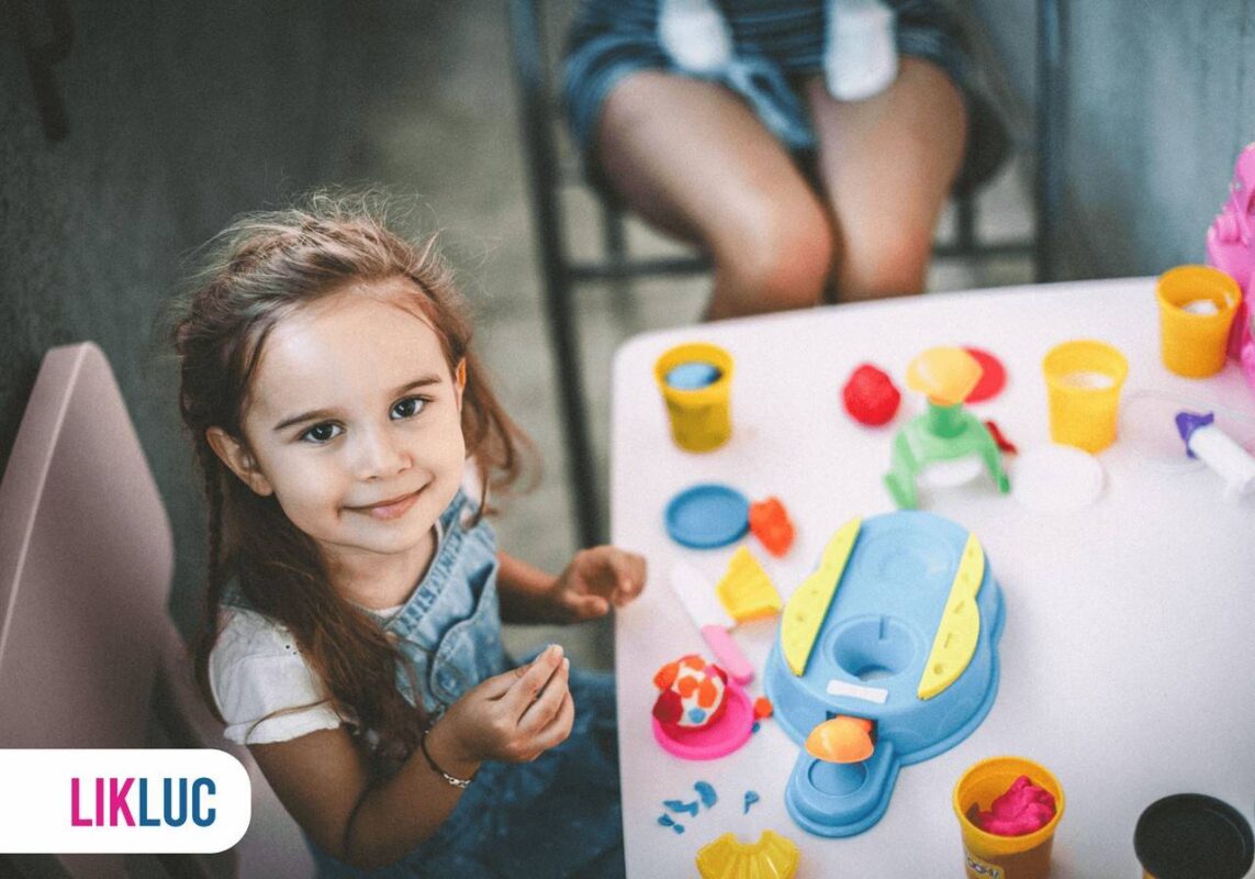Criança sentada em cadeira, com vários brinquedos sobre a mesa inclusive massinha de modelar