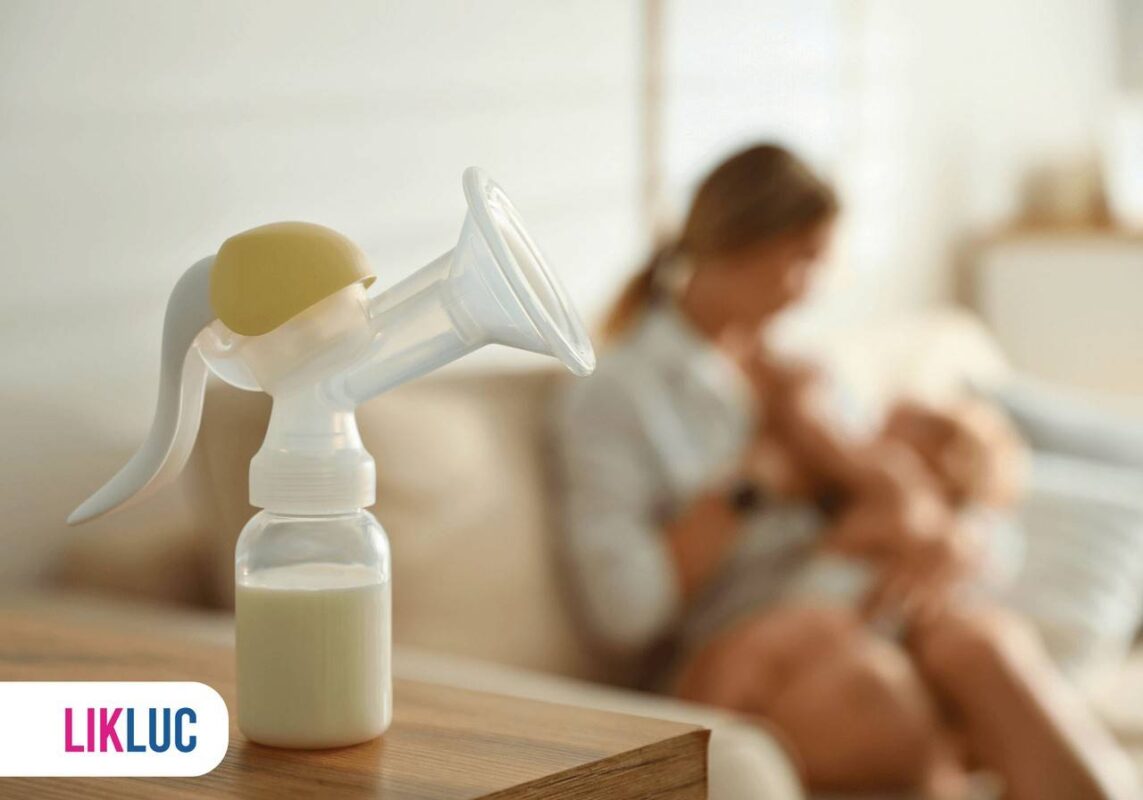 Bomba de tirar leite em cima de mesa em madeira, ao fundo mãe amamentando bebê no sofá