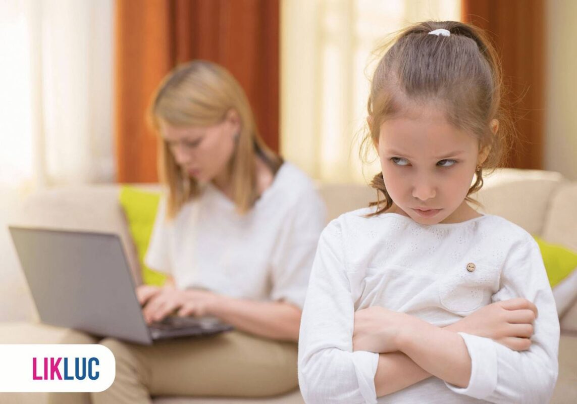 Criança brava de braços cruzados, ao fundo mãe sentada em sofá com notebook em mãos