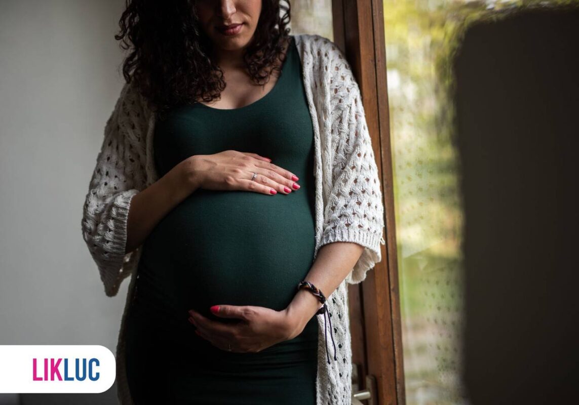 Gestante em pé com as duas mãos sobre a barriga ao lado de porta de vidro
