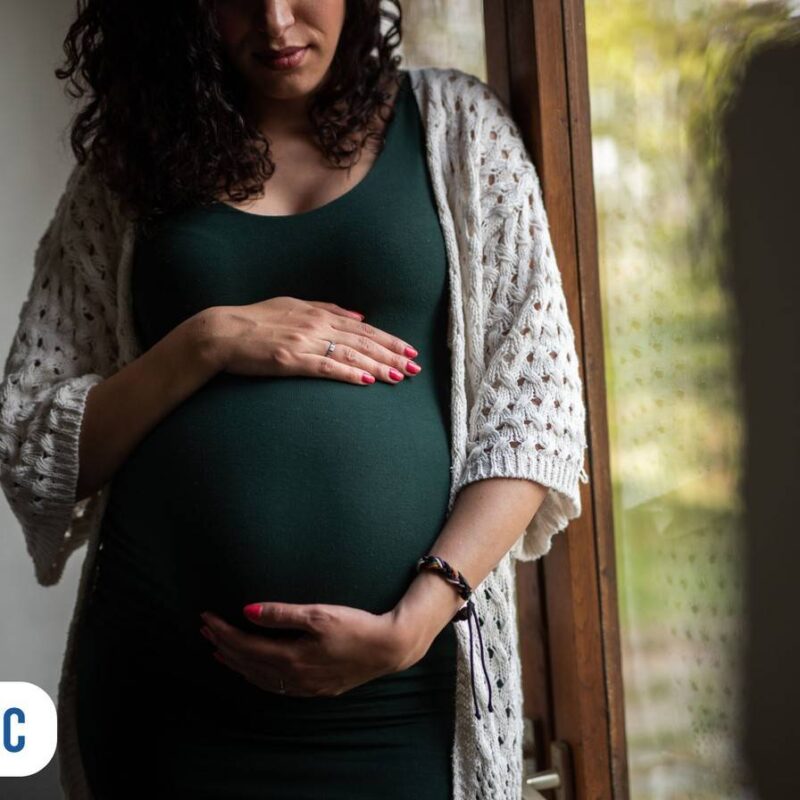Gestante em pé com as duas mãos sobre a barriga ao lado de porta de vidro