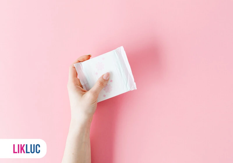 Mão segurando um pacote de absorventes sobre fundo rosa.