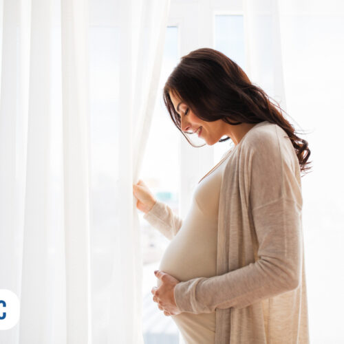 Mulher grávida olhando pela janela com cortinas brancas.
