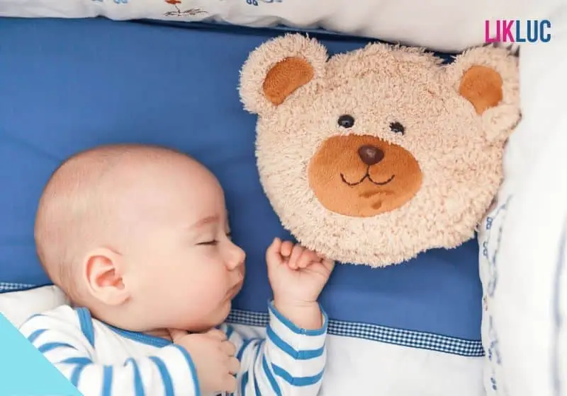 Bebê dormindo de lado no berço com travesseiro e um ursinho de pelúcia
