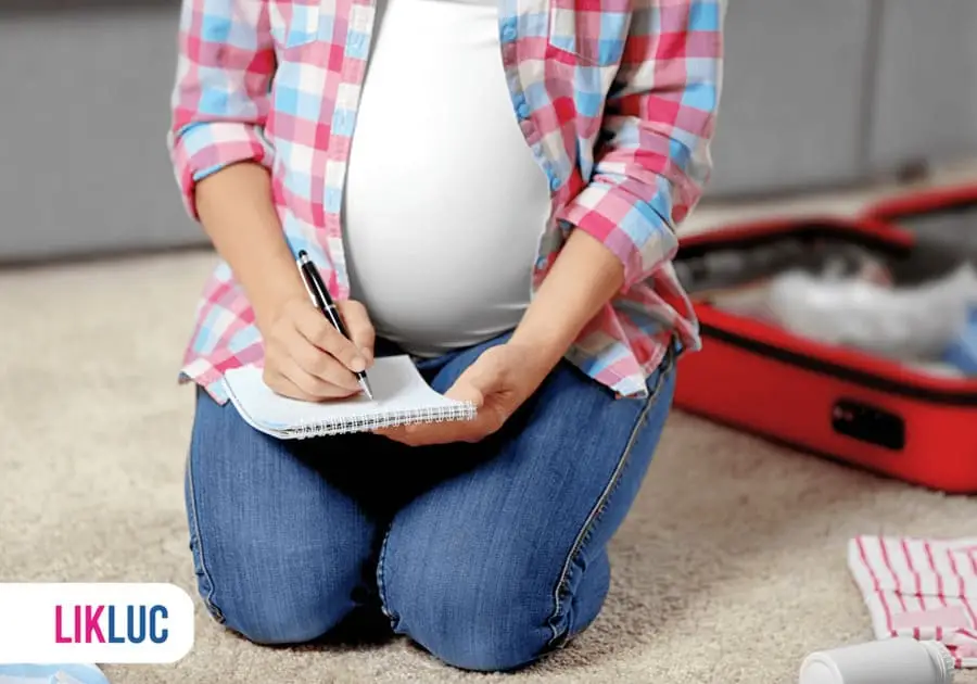 Gestante sentada no chão ao lado da sua mala de maternidade escrevendo a lista do enxoval