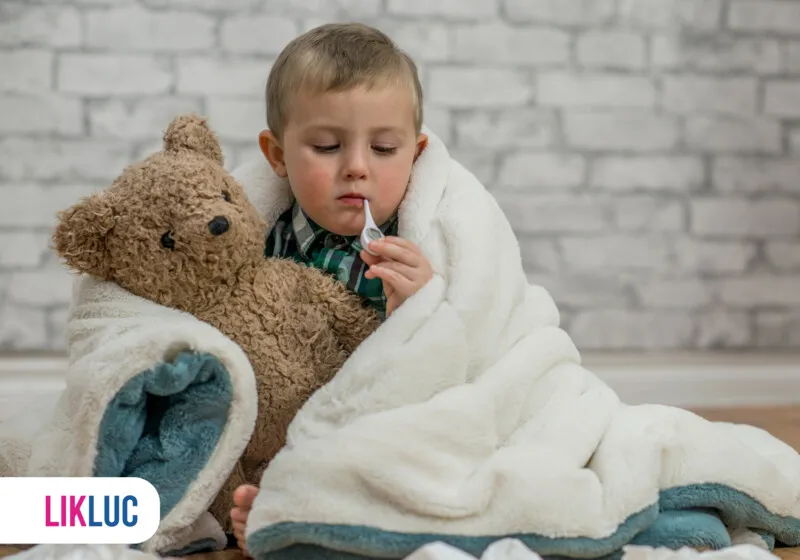 Criança com cobertor e urso de pelúcia medindo temperatura.