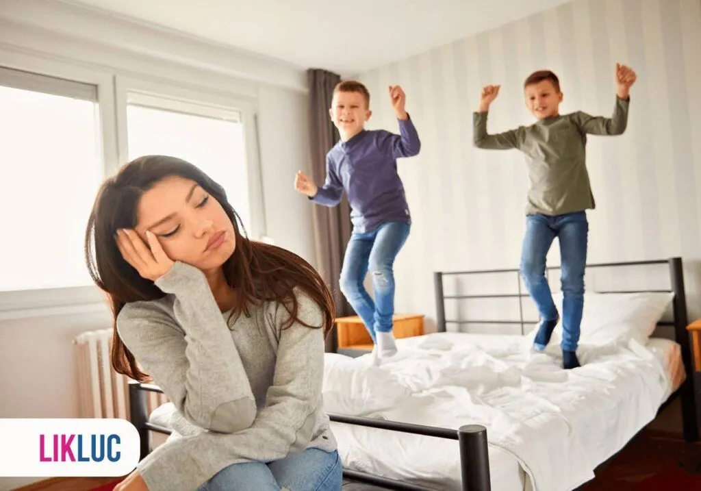 Duas crianças pulando na cama e a mãe sentada cansada