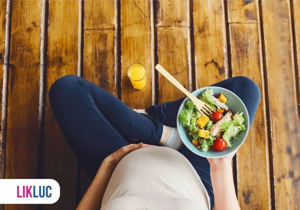 Gestante com uma mão na barriga e na outra segurando um prato com salada, ao lado um copo com suco de laranja